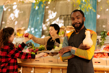 Male vendor selling wholesome, sustainably produced produce while positioned close to food market. African american farmer wearing an apron is holding fresh organic squash and facing the camera.