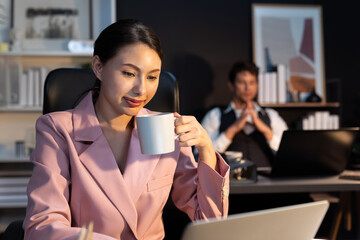 Focusing on sitting woman coworker drinking tasty coffee on working front desk while waiting email business project report at late night time with blurry man background working on laptop. Postulate.