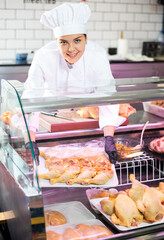 Smiling skilled young female butcher working behind counter in butchery, putting tray with fresh raw chicken leg quarters into glass showcase