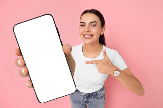 Happy woman pointing at mobile phone with blank screen on pink background. Mockup for design