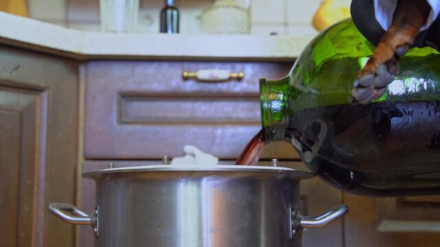 A hand is carefully pouring the liquid from a green bottle into a shiny stainless steel pot in a cozy kitchen