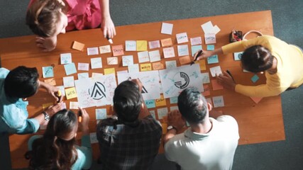 Top down aerial view of skilled designer hold logo while discuss with team. Aerial business group working together sharing idea about logo graphic design by using sticky notes and tablet. Symposium.