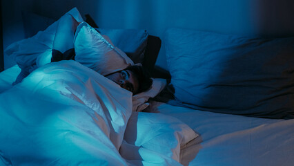 Pensive man trouble sleeping disturbed in bed with soft pillows under soothing dim blue light.
