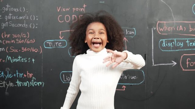 African girl smiling and waving at camera while standing at board with engineering prompt or coding, programing system written in STEM classroom. Child looking at camera while greeting. Erudition.