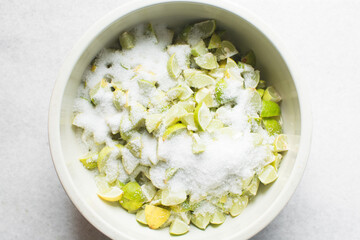 Overhead view of lime slices and sugar being mixed together in a white bowl, process of making lime cheong or lime syrup