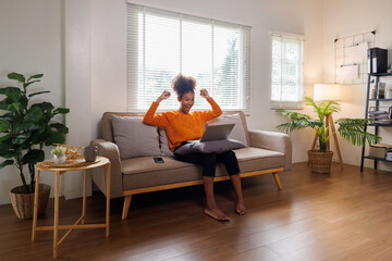 Image of cheerful excited happy african american woman afro hair,  using laptop while sitting on chair in living room