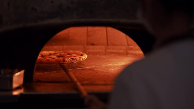 A professional chef wearing his uniform, skillfully maneuvers a pizza with a crispy golden onto the stone inside a wood fired oven. Traditional cooking, rustic charm to this Italian culinary delight.