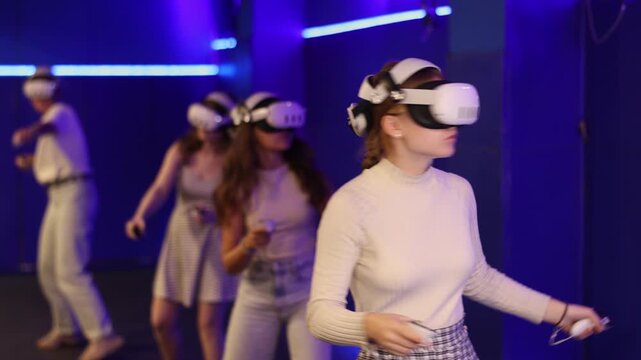 Group of excited young girls standing with handheld controllers in hands engaged in virtual world experiencing through VR glasses covering face. Concept of immersion and interactivity in game