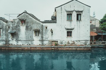 A weathered white building with windows stands beside a still canal in Suzhou, China.