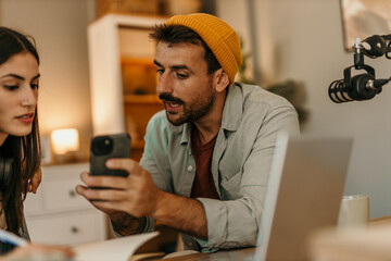 Man and woman podcasting with professional equipment in a cozy living room, man showing something...
