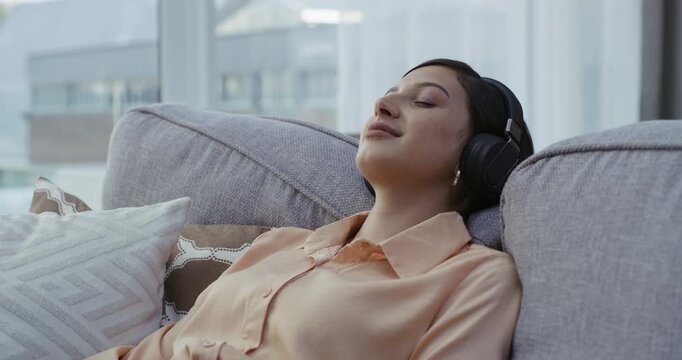 Wearing headphones, listening to relaxing music from phone and enjoying a calm, zen and peaceful day at home. Happy and relaxed woman sitting on a living room sofa and meditating with binaural songs