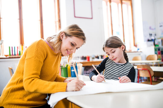 Hardworking teacher learning with young schoolgirl in classroom, sitting by desk.