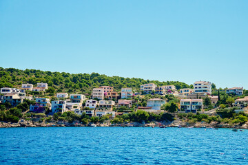 Fototapeta premium Seaside town with colorful coastal houses on hillside with boats docked along clear blue waters. Residential apartments for summer vacation on Adriatic Sea in Rogoznica, Croatia