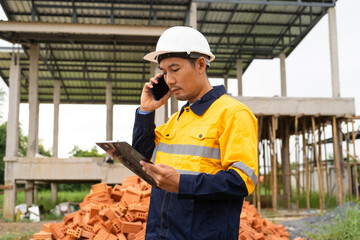 A male architect wearing a hard hat and safety vest checks his laptop while holding a clipboard...