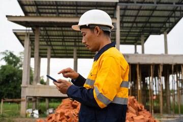 A male architect wearing a hard hat and safety vest checks his laptop while holding a clipboard with house plan papers. He reviews blueprints, construction documents,house structure details on-site.