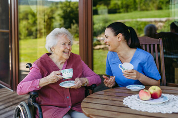 Female caregiver spending time with elderly woman, drinking coffee and talking. Nurse and senior...
