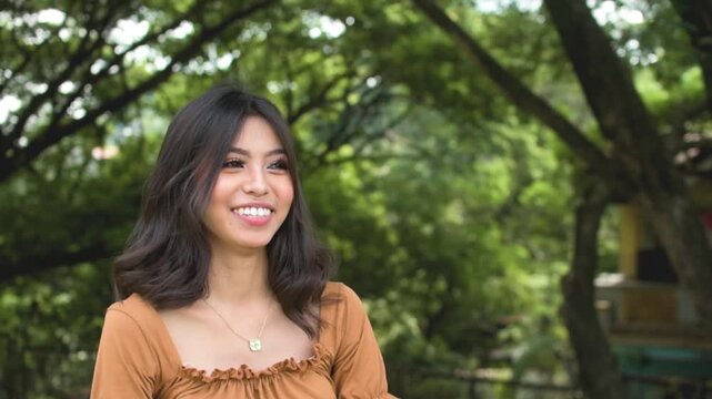 A young and shy asian woman blushes after received flattering compliments. Regains composure shortly and smiles at the camera. Outdoor park scene.