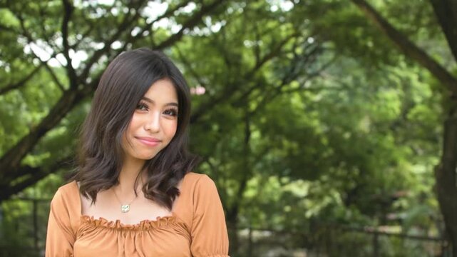 A young teenage asian woman trying to look cute for someone, smiling trying to get attention. Outdoor park scene. Copy space on the right. Slowed down 50 percent.