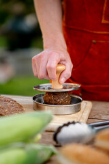Man in apron preparing meat for hamburgers, standing outdoors. Using burger patty press to shape groud meat.