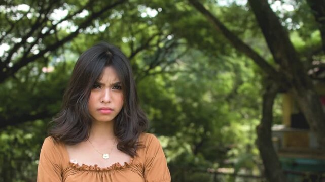 A young teenage asian woman looks annoyed and upset, refusing to budge. Outdoor park scene. Copy space on the right. Slowed down 50 percent.