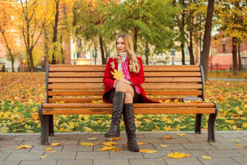 A girl in a red coat sits on a bench in an autumn park. She holds yellow leaves in her hand.