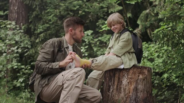 Caucasian tween boy sitting on tree stump in forest while his father taping his injured ankle