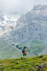 Fototapeta premium hiking in the mountains. A little boy with a backpack walks along a path against a background of mountains and clouds. Active healthy lifestyle on weekend hike journey.