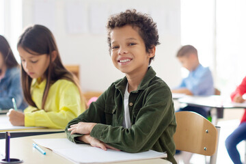 Obraz premium Smiling schoolboy sitting at desk in classroom and smiling, classmates writing in copybooks on the background. Education, learning and people concept