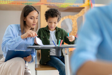 Cheerful young woman teacher explaining to latin male student grammar rules, sitting near boy desk,...