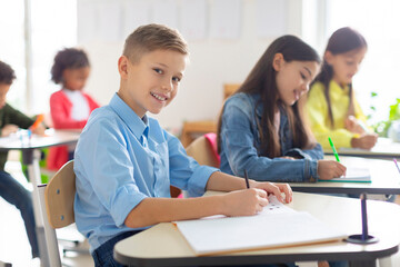 European schoolboy sitting at his desk in classroom and smiling at camera during lesson in school