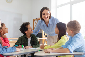 Kids sitting at desks in classroom and listening their teacher, woman explaining something interesting to pupils, studying at school