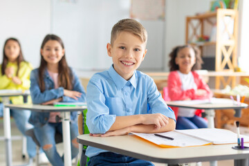 Fototapeta premium Happy European schoolboy sitting at school desk and smiling at camera with diverse classmates on background. Smart kids and education concept