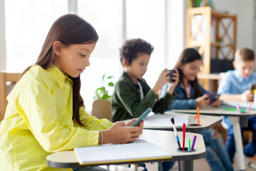Multiethnic kids pupils having break and using their smartphones, sitting at desks in classroom, playing games, focus on girl