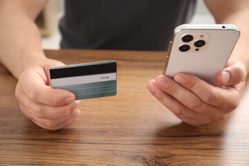 Man with credit card and smartphone at wooden table indoors, closeup