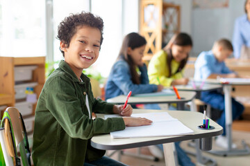Happy Latin boy sitting at desk in classroom, preparing for exam test or writing project, looking at camera and smiling