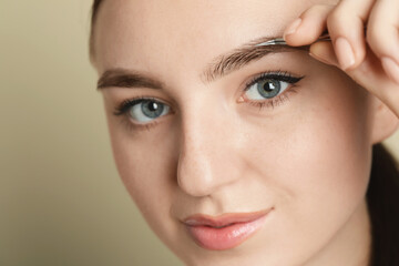 Fototapeta premium Young woman plucking eyebrow with tweezers on beige background, closeup