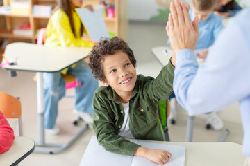 Female teacher giving high five to Latin schoolboy sitting at desk, supporting kid, achievement,...