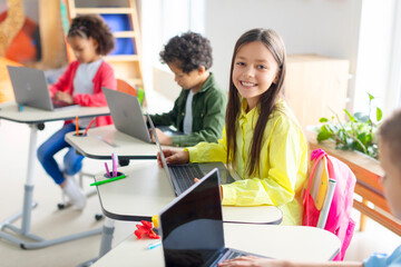 Cheerful schoolgirl using laptop computer, learning online with classmates sitting at desks in modern classroom. Technology and education concept
