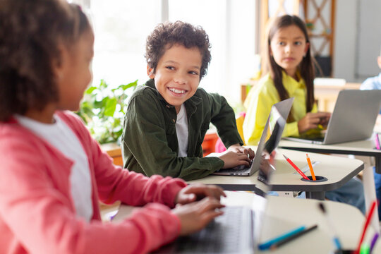 Latin schoolboy talking to girl while having tech lesson, children using laptops. Kid actively discussing and collaborating with friend
