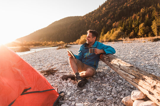 Smiling man holding tablet PC and leaning on log near riverside