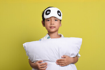 Portrait of cute Asian boy wearing pajamas and sleep mask holding pillow isolated on yellow background. Healthy sleep concept. Copy space