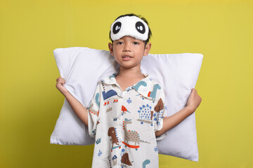 Portrait of cheerful Asian boy wearing pajamas and sleep mask holding pillow isolated on yellow background. Healthy sleep concept. Copy space