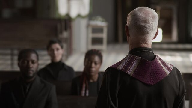 Back view of senior pastor reading prayer in front of three grieving people sitting on wooden benches during funeral service in church