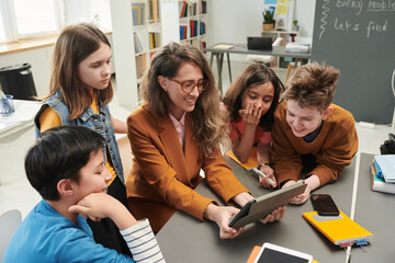 High angle view at smiling female teacher using tablet with diverse group of kids watching video...