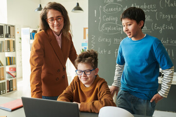 Portrait of smiling female teacher and two young boys looking at laptop screen in school classroom...
