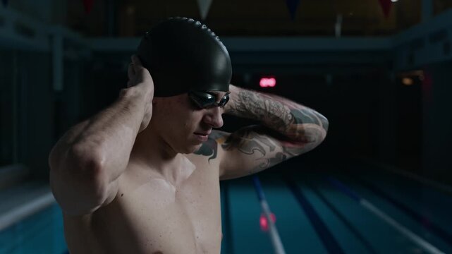 Tilt up medium shot of Caucasian male swimmer in indoor pool putting on black swimming cap while getting ready for race