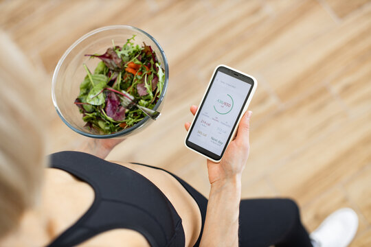 Woman in fitness clothing tracking daily calorie intake on smartphone while holding bowl of fresh salad. Concept of healthy eating, fitness, nutrition, and technology.
