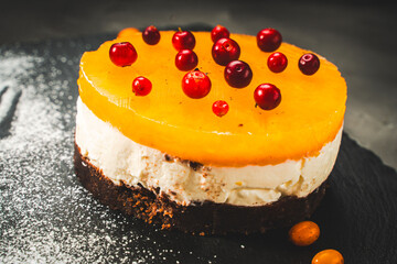 A cake or dessert made of mango or sea buckthorn jelly, layered with cottage cheese cream and chocolate cookies, decorated with lingonberries and powdered sugar on a black plate on a dark background.