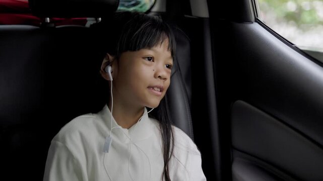 An 8+ year old Asian elementary school girl with a cute face, wearing a white long-sleeve shirt, is traveling in a car she is happily listening to her favorite music with her earphones on, Smiling fac