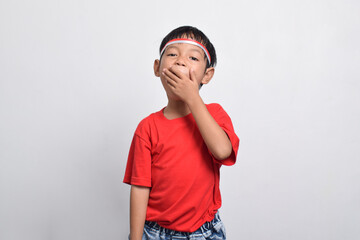 A cheerful Indonesian boy in a red shirt looks surprised while covering his mouth, isolated on a white background. Indonesian Independence Day concept.